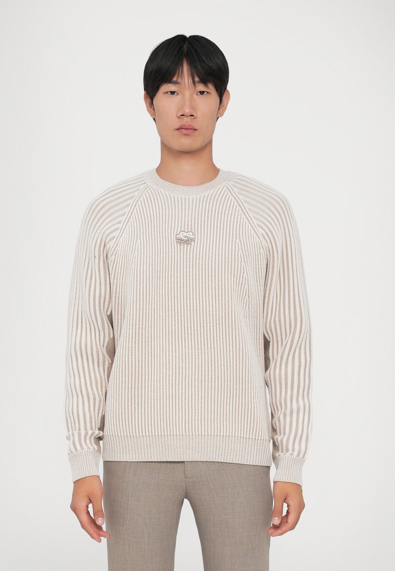Jonge man in een beige en witte gestreepte gebreide trui met een klein sluitingdetail en lichtbruine broek tegen een effen achtergrond.