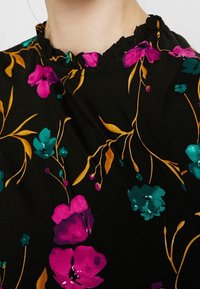 Schwarzes Blumenshirt mit einem Rüschenausschnitt. Ausgestattet mit lebhaften rosa und türkisfarbenen Blumen, goldenen Blättern und einer weichen Stoffstruktur.
