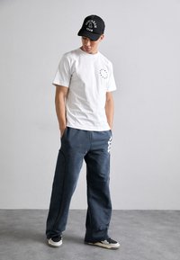 Hvit t-skjorte med rundt logo på brystet, marineblå joggebukser med sideakenter, og svart caps med hvit tekst. Uformelt fottøy fullfører antrekket.