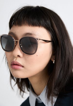 Solbriller med mørke linser og klare brune stel, med metalliske detaljer på stængerne. Kvinde med kort, mørkt hår iført en skjorte med krave.