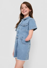 Robe salopette en denim bleu clair, manches courtes, boutonnière sur le devant, deux poches poitrine, ourlet effiloché. Elle présente un accent de logo sur la manche.