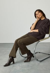 Frau mit langen dunklen Haaren, die einen lockeren dunklen Sweatshirt, braune Hosen und spitze Knöchelstiefel trägt, sitzt auf einem modernen Stuhl aus Chrom.