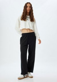 Witte cropped sweatshirt met zwarte tekst, gecombineerd met losse zwarte broek met blauwe zijstrepen en witte sneakers met zwarte accenten.