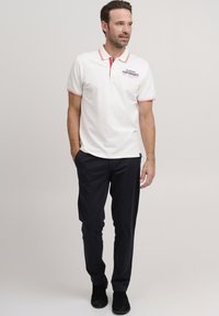 Hvit polo skjorte med røde detaljer, med en logo på brystet. Kombinert med svarte bukser og svarte sko, fremhever antrekket uformell bekledning.