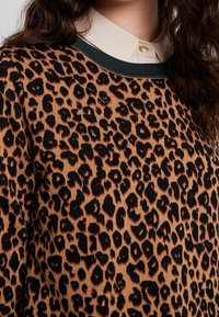 Pull en imprimé léopard beige chaud avec des taches noires. Il présente un col côtelé vert foncé et une superposition de col clair en dessous.