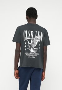 Pilka medvilninė marškinėliai su dideliu baltu erelio grafiku ir tekstu ant nugaros, dekoruoti penkiomis žvaigždėmis. Trumpomis rankovėmis, laisvas kirpimas.