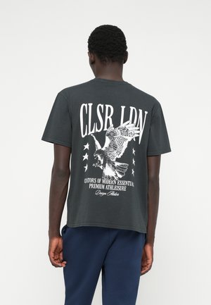 Sivý bavlnený tričko s veľkým bielym grafickým vyobrazením orla a textom na chrbte, s piatimi hviezdami zdôrazňujúcimi dizajn. Krátke rukávy, voľný strih.