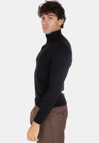Maglione a collo alto nero, aderente, con maniche lunghe e polsini a costine, abbinato a pantaloni marroni. Tessuto a maglia liscio con una silhouette pulita.