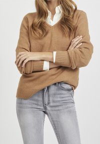 Bruine v-hals trui van zachte brei met geribbelde manchetten en onderkant, gedragen over een witte blouse met kraag. Gecombineerd met lichtgrijze slimfit jeans.