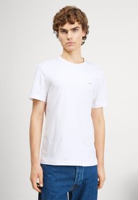 White cotton t-shirt with short sleeves, round neckline, and a small brand logo on the left chest. Paired with blue denim jeans.