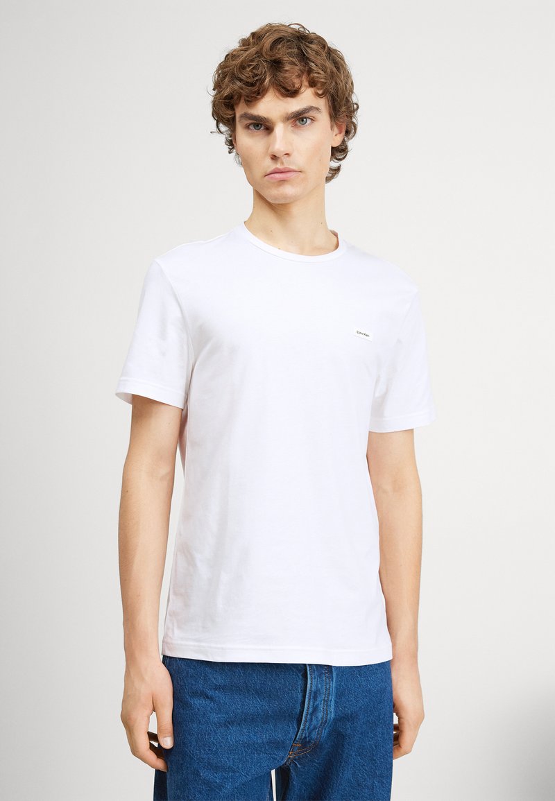 White cotton t-shirt with short sleeves, round neckline, and a small brand logo on the left chest. Paired with blue denim jeans.