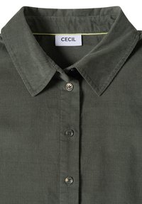 Chemise en velours côtelé vert foncé avec col pointu et boutons sur le devant, affichant une étiquette blanche avec le nom de la marque Cecil.