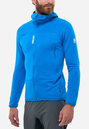 Homme portant un sweat à capuche zippé bleu vif avec deux poches poitrine zippées et un pantalon gris, debout devant un fond blanc.