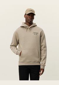 Sudadera beige con bolsillo frontal y capucha con cordón. Presenta un logo bordado en el pecho. Combinada con una gorra a juego.