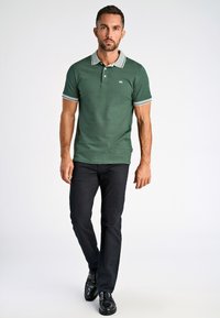 Camisa polo verde con cuello y acentos en las mangas grises, combinada con pantalones negros y zapatos negros. El modelo está de pie frente a un fondo neutro.