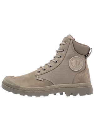 Beżowy, wysokiej klasy, wytrzymały but za kostkę z metalowymi oczkami, teksturowanymi bokami ze tkaniny, gumową podeszwą oraz okrągłą naszywką z logo Palladium w pobliżu kostki.