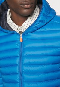 Giacca imbottita blu con chiusura a zip e un tira arancione, superficie testurizzata e cappuccio. Camicia a righe visibile sotto.