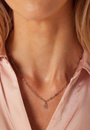 Collier en chaîne argentée avec des maillons allongés et un petit pendentif en forme de lettre "E". Porté avec une chemise rose clair, mettant en valeur son design et son éclat.