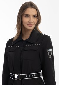 Geacă utilitară neagră cu studs argintii pe umeri, două buzunare frontale și o curea asortată cu cuvântul "IUBIRE". Accent de lanț pe mânecă.