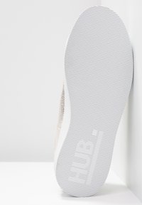 Semelle de chaussure vue de dessous, présentant un caoutchouc blanc texturé avec des rainures horizontales et une empreinte de logo. La partie supérieure a un design clair et perforé.