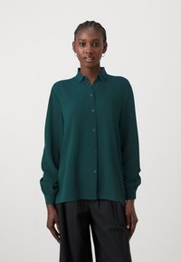 Cămașă verde cu nasturi dintr-o țesătură ușoară, cu guler, mâneci lungi și manșete cu nasturi, asortată cu pantaloni largi închiși la culoare.