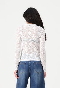 Hvit blonde topp med lange ermer, blomsterdesign og ribbestruktur. Har rund hals og tettsittende silhuett, kombinert med blå jeans.