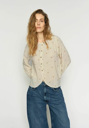 Donna con capelli lunghi e mossi che indossa una camicetta color crema con piccoli motivi floreali e jeans blu larghi, in piedi davanti a uno sfondo semplice.