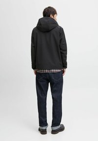 Veste imperméable noire avec capuche, portée par-dessus une chemise à carreaux, associée à un jean en denim noir et des chaussures marron, vue de dos.