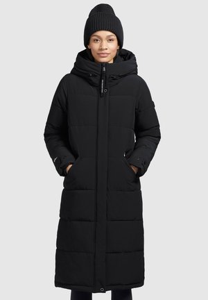 Black long puffer coat with a hood, featuring a quilted design, side pockets, and adjustable cuff details. Paired with a black knit beanie.