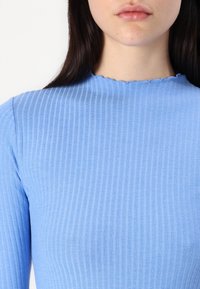 Top blu ribbed con scollo scallop e maniche lunghe, realizzato in un tessuto testurizzato che aggiunge interesse visivo.