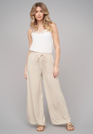 Junge Frau trägt weißes Spitzen-Camisole, beige weite Hosen mit Kordelzug und flache Sandalen, steht vor schlichtem grauem Hintergrund.