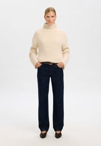 Smėlinio spalvos vilnonis golfas su gofruotomis rankovėmis, derinamas su tamsiai mėlynomis tiesiomis kelnėmis ir tamsiais zomšos aukštakulniais. Paprastas ir švarus dizainas.