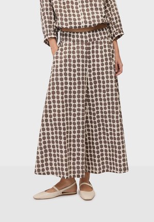 Femme portant un pantalon large beige à gros pois marron et des chaussures plates beiges à brides à la cheville, debout les mains dans les poches.