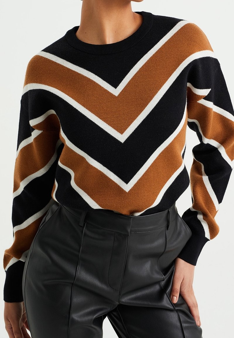 Personne portant un pull à motif chevron noir, marron et blanc, rentré dans un pantalon en cuir noir taille haute, main posée sur la hanche.