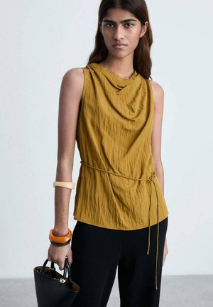 Cowl-neck, sleeveless top in mustard yellow, textured fabric. Paired with black pants and a black handbag, featuring orange and clear bangles.