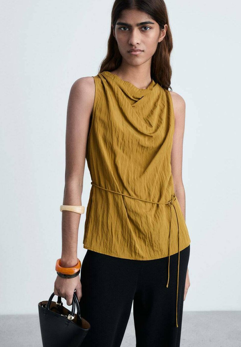 Top fără mâneci, cu guler cowl, de culoare muștar, fabricat dintr-un material texturat. Asortat cu pantaloni negri și o geantă neagră, având brățări portocalii și transparente.