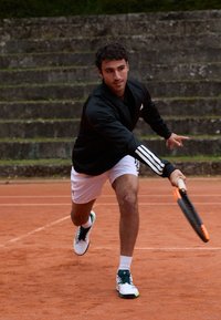 Jovem a avançar num campo de ténis de argila, vestindo uma jaqueta preta, calções brancos, segurando uma raquete pronto para bater na bola.