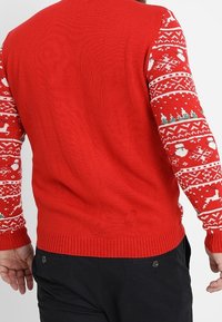 Pull rouge avec manches à motifs comprenant des bonhommes de neige blancs, des arbres et des rennes. Matière tricotée, poignets et ourlet côtelés. Design classique.