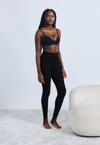 Colanți negri cu talie înaltă, textură moale și detaliu deschis la gleznă, asortați cu un bralette negru. Fundalul este gri deschis.