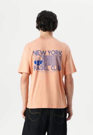 Perzik kleurige katoenen t-shirt met korte mouwen en een relaxte pasvorm. Heeft een grafische print in het blauw met de tekst "NEW YORK PADEL CLUB" op de achterkant.
