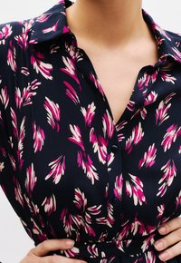 Blouse bleu marine avec un motif floral rose et crème, patte de boutonnage et col. Tissu doux et détail de taille froncée.
