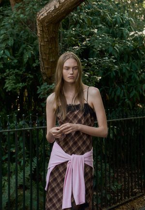 Jeune femme aux longs cheveux portant une robe à carreaux marron et un pull rose noué autour de la taille, debout près d’un arbre et d’une clôture en fer dans un parc verdoyant.