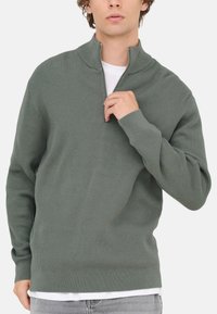 Pullover verde a coste con colletto alto e design con mezzo zip, caratterizzato da maniche lunghe e una vestibilità rilassata, indossato sopra una camicia bianca.