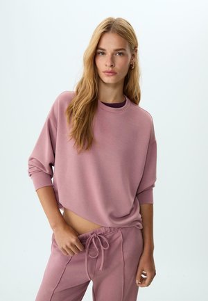 Giovane donna con lunghi capelli biondi, vestita con una felpa over size color malva e pantaloni abbinati, in posa su uno sfondo chiaro e semplice.