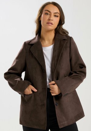 Femme portant un blazer marron foncé boutonné sur une chemise blanche, une main dans la poche et l'autre ajustant le devant.