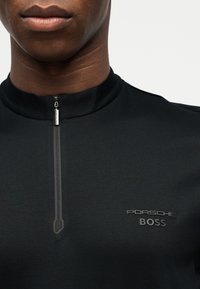 Κοντινό πλάνο ενός ατόμου που φοράει μαύρο επάνω μέρος με κοντό φερμουάρ και το λογότυπο "Porsche BOSS" στο στήθος.