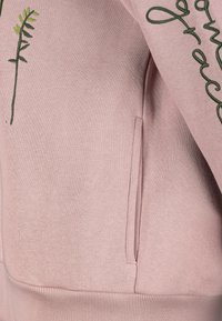 Rosa Sweatshirt mit grün gesticktem Blattmotiv und dem Schriftzug "grace" in Schreibschrift. Mit seitlicher Tasche und gerippten Bündchen.