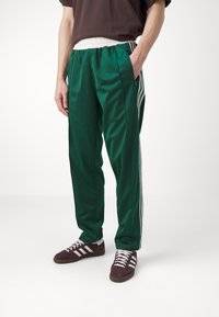 Gröna satinbyxor med vita sidosträck, elastisk midja och sidofickor, i kombination med mörka sneakers med vita ränder.