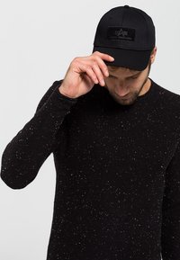 Crna bejzbol kapa s teksturiranim crnim pečatom s logotipom "Alpha Industries"; model nosi crni džemper s pjegicama.