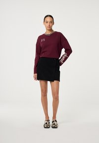 Bordeaux langemouwshirt met logodetail, gecombineerd met een zwarte corduroy rok met een asymmetrische zoom. Leoprint instap schoenen.